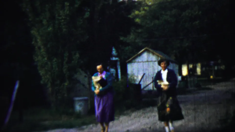 Two women in 1950s attire walk along a dirt path in Chadron, Nebraska, near trees, a shed, and residential buildings.