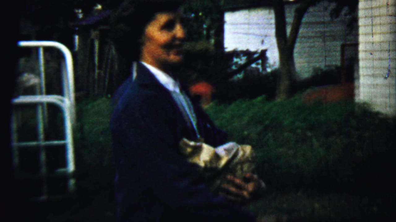 A woman in dark 1950s clothing holds a wrapped bundle near a white clapboard house and greenery in Chadron, Nebraska.