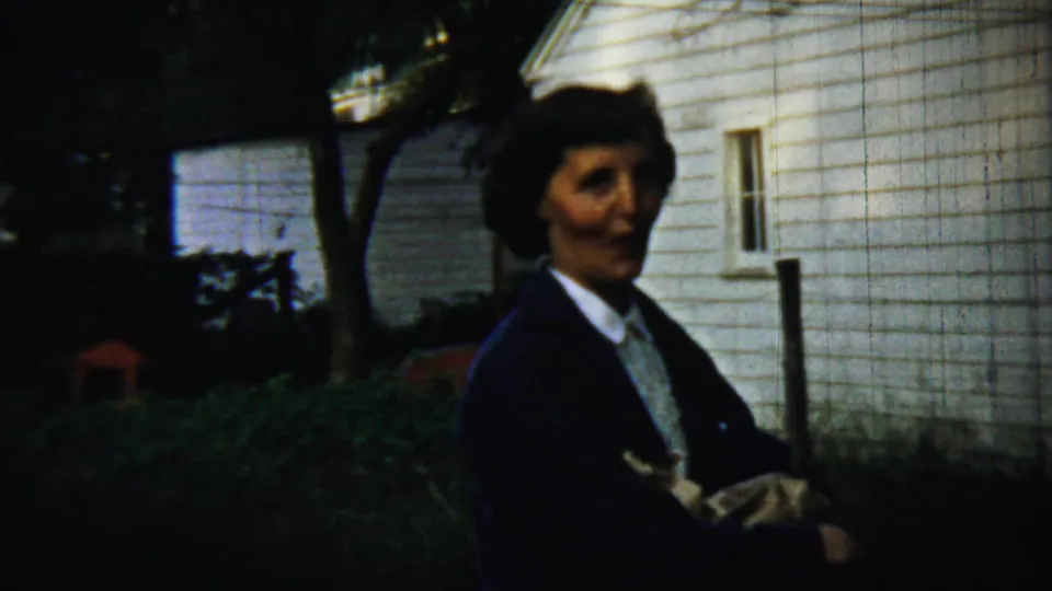 A woman in mid-century dress and jacket smiles near white clapboard siding while holding a wrapped bundle outdoors.