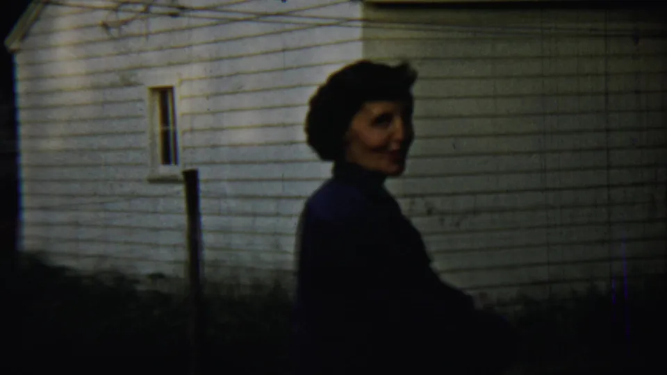 A woman in profile passes beside a white clapboard house in a shaded Chadron residential yard.