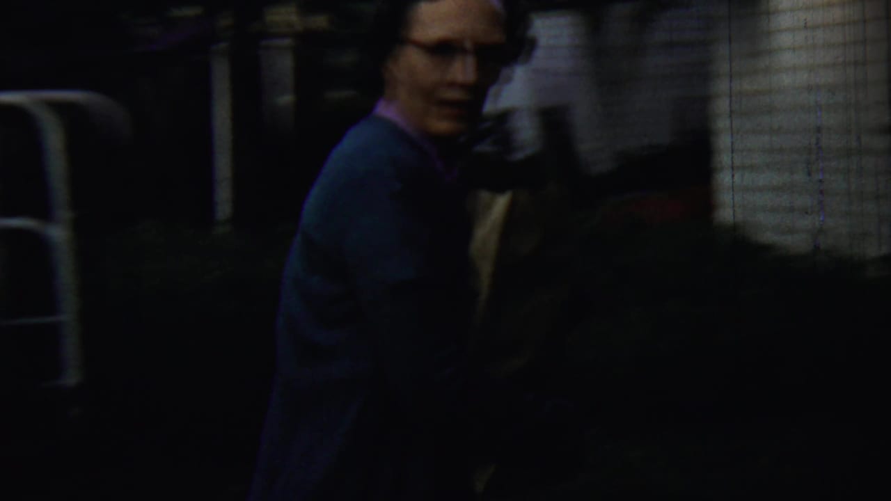 Woman in a blue cardigan stands near a dark yard beside a white-sided house.