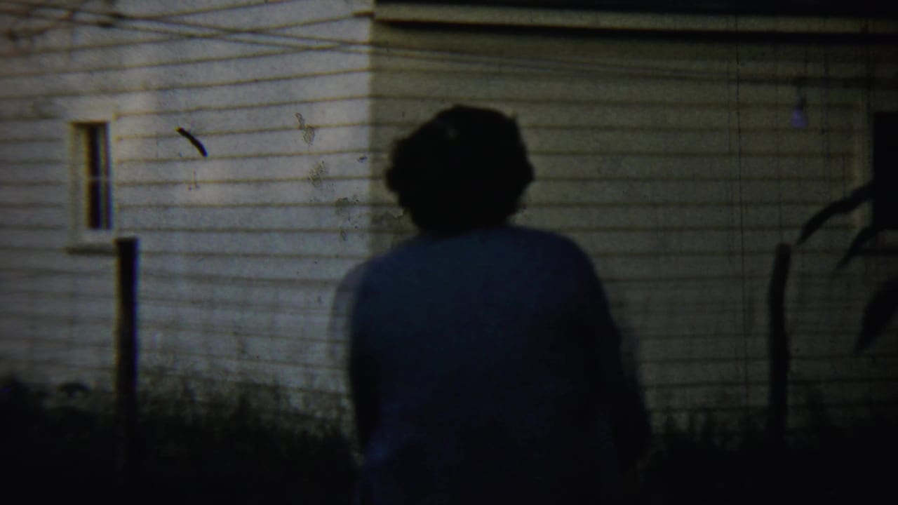 A woman walks away from the camera beside a white-sided house in dusk light, consistent with the 1959 Chadron home-movie metadata.