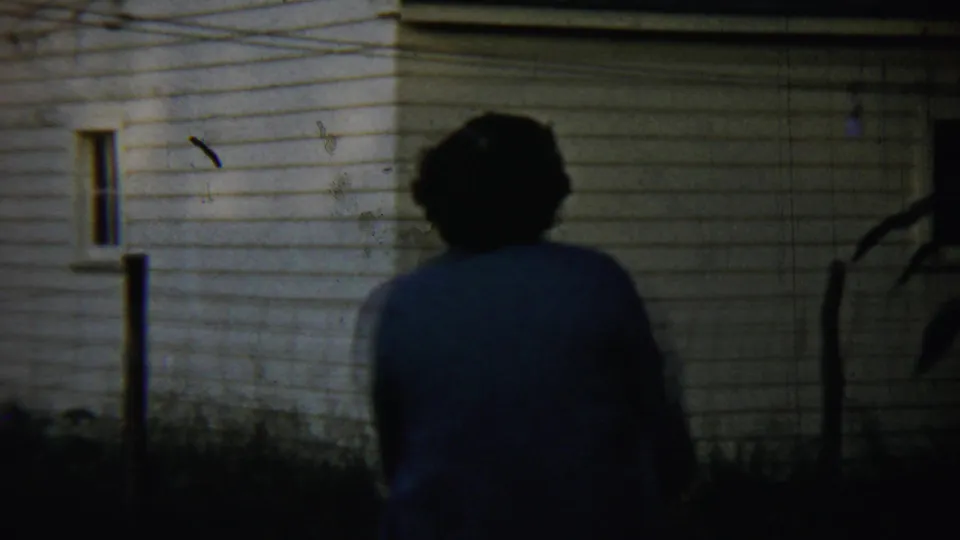 A woman walks away from the camera beside a white-sided house in dusk light, consistent with the 1959 Chadron home-movie metadata.