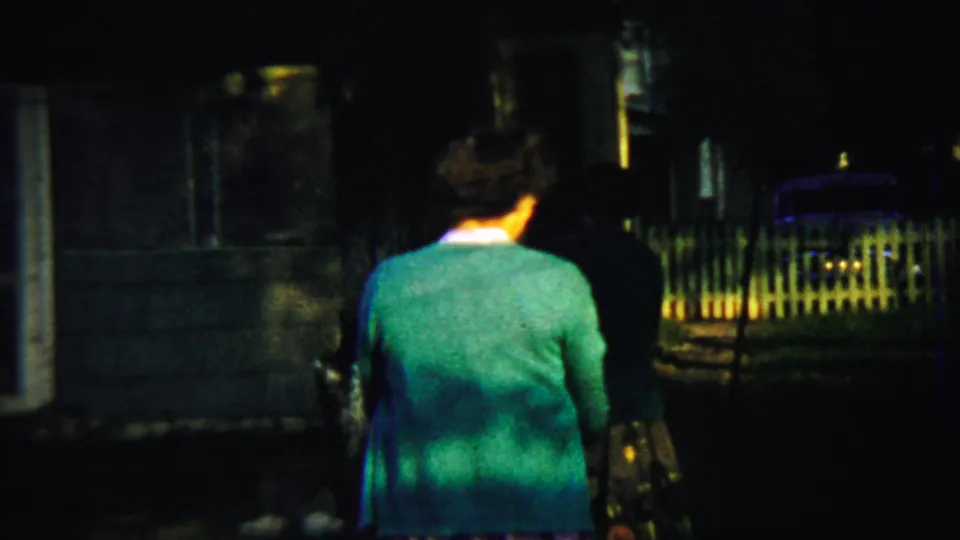 The woman walks through a shadowed yard toward a porch area, with a picket fence and parked car visible in the background.