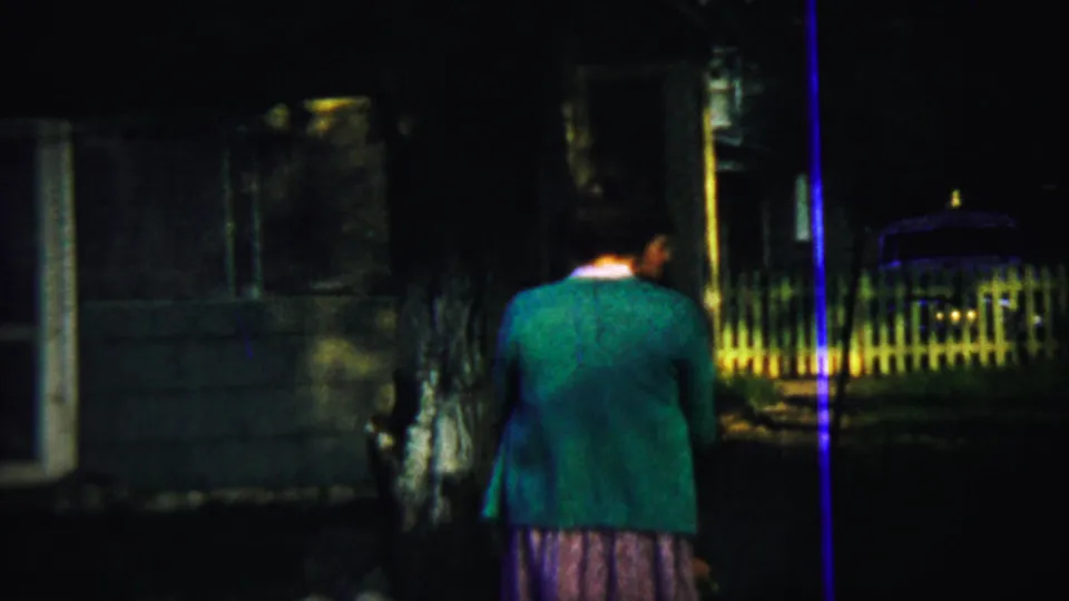 At the end of the 1959 Chadron clip, the woman continues through the dim yard with a fence, posts, and parked car behind her.