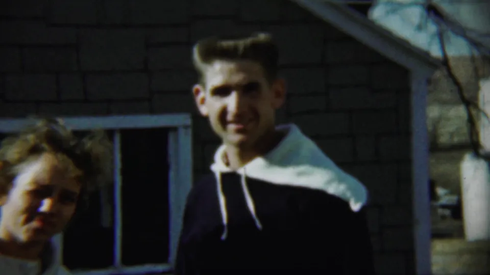 A young man smiles and a young woman leans forward outside a brick house in 1959 Chadron, Nebraska home-movie footage.