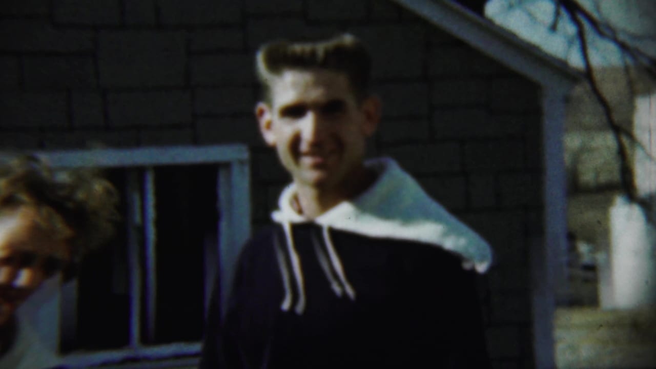 Young man smiling outside a brick house while a woman is partly visible at the edge.