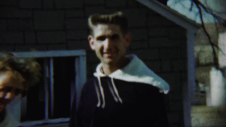 In Chadron, Nebraska, 1959, a young man smiles near a brick house as a woman appears cropped at the left edge.