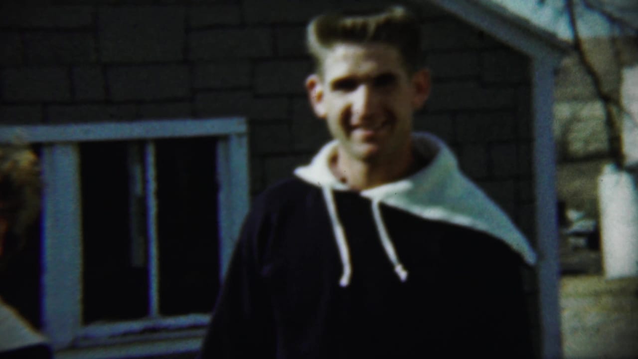 Young man in a dark hooded top stands outside a brick house with bare branches nearby.