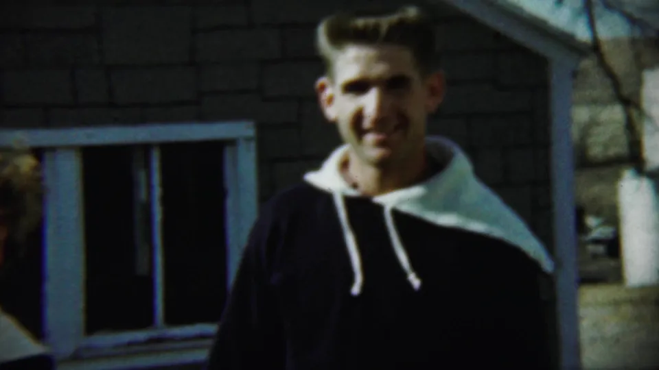 In the 1959 Chadron home movie, a young man faces the camera outside a brick house with a window and bare branches behind him.