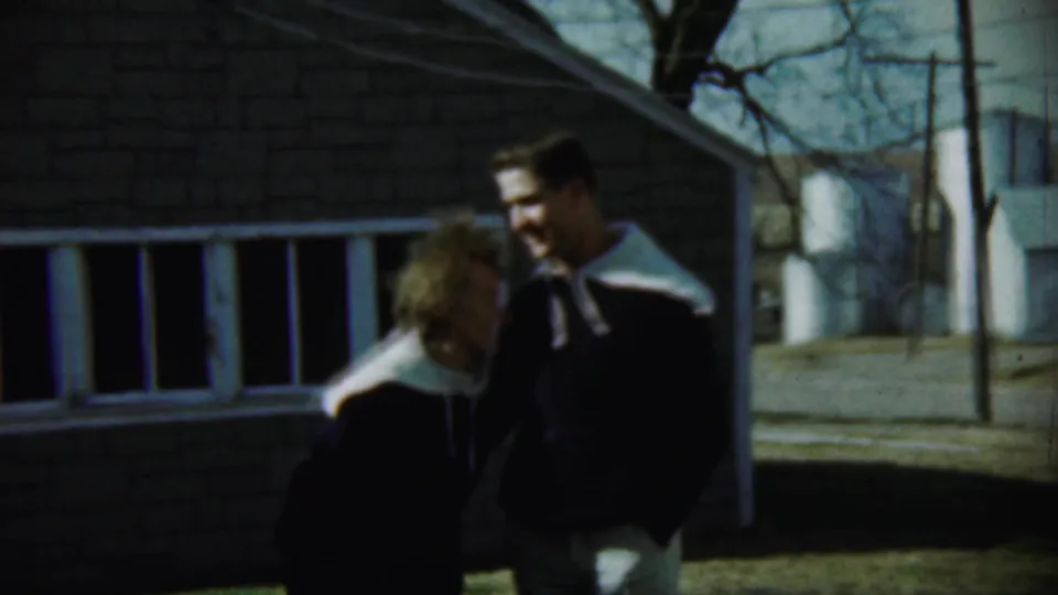 A woman embraces or leans against the tall young man outside a brick house in Chadron, Nebraska, in the 1959 home movie.