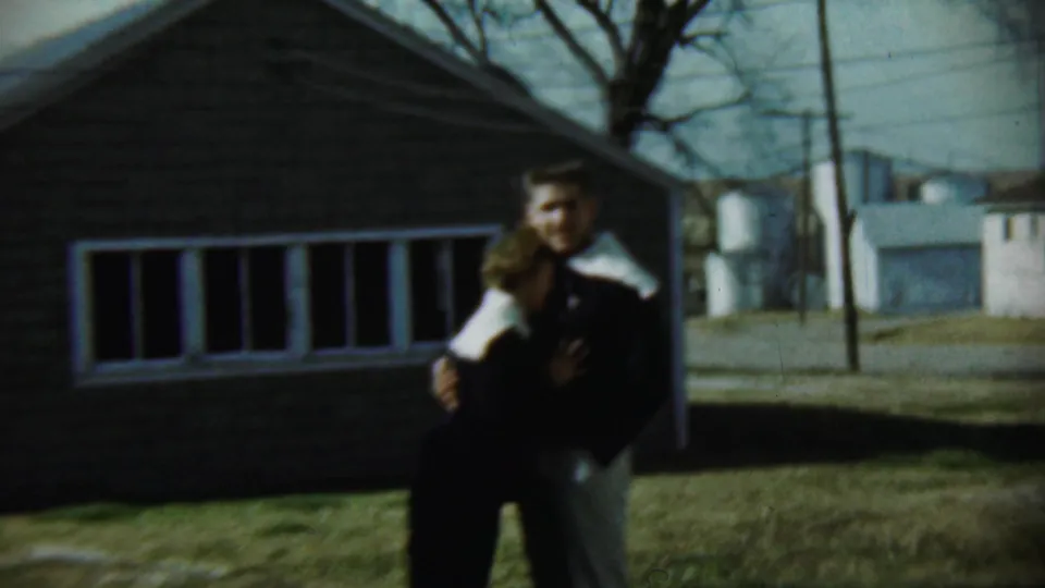 In 1959 Chadron, Nebraska footage, a young couple stands close together outside a brick house on a grassy lawn.