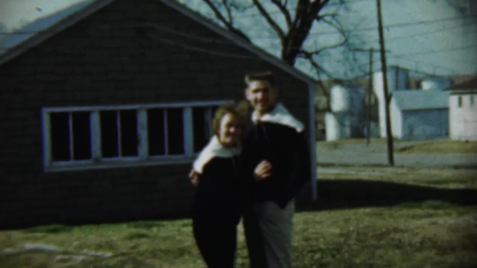 The couple faces the camera in a 1959 Chadron, Nebraska home-movie frame, standing arm in arm beside a brick house.