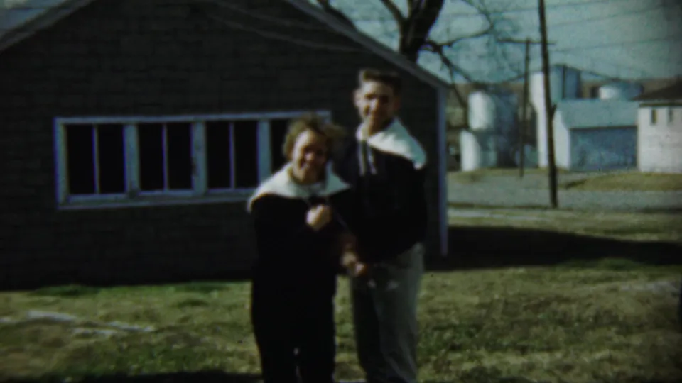 A smiling woman and a man stand together on a lawn in Chadron, Nebraska, with a brick house and rural backdrop visible.