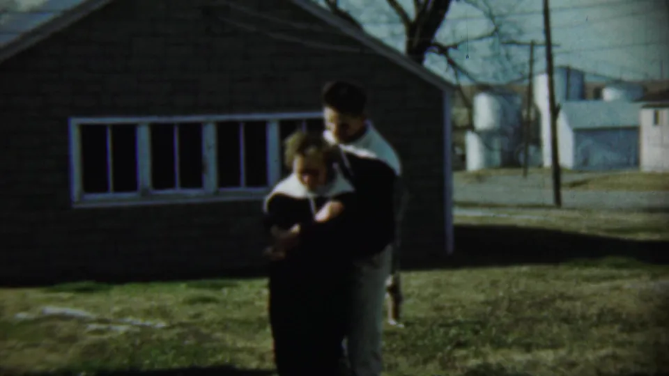 The couple stands close together outside the brick house in the final 1959 Chadron, Nebraska frame, with rural buildings in the background.