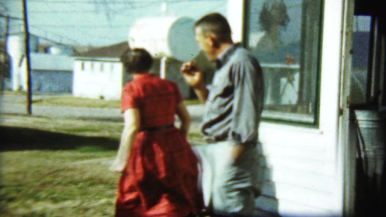 Woman in a red dress walks near a man smoking beside a white house.