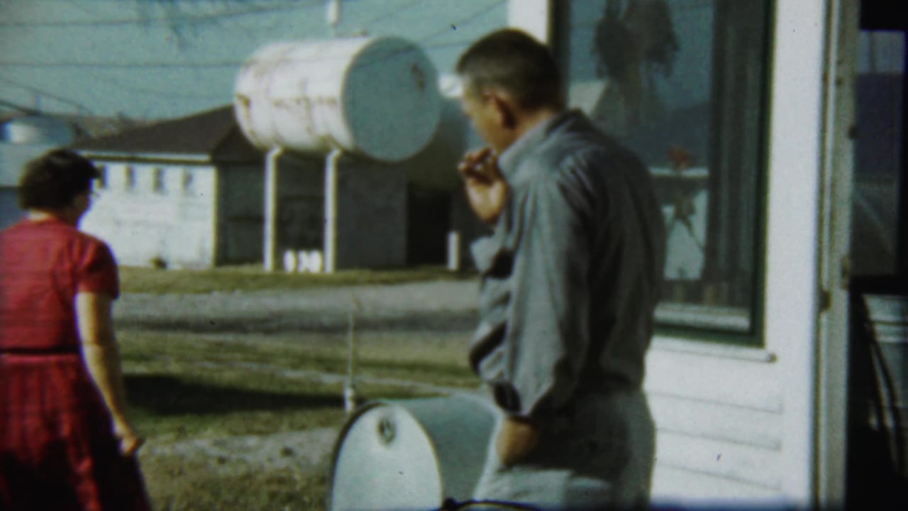 Woman in red dress walks away while a man smokes near the house corner.