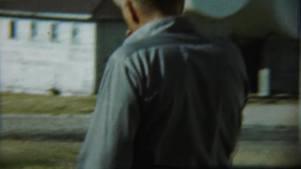 A closer handheld frame shows the man's back in a gray shirt, with the yard and light-colored building beyond in Chadron, Nebraska footage.
