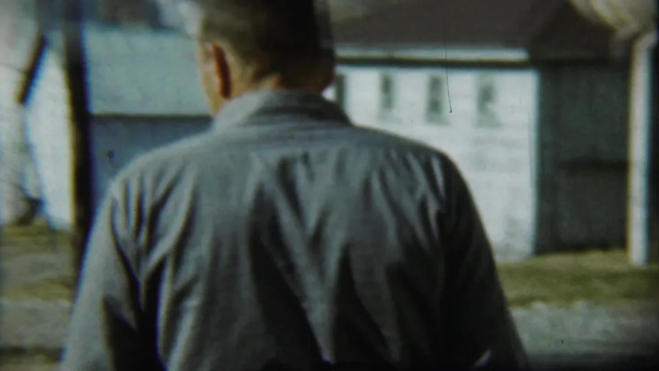 The final frame holds on the man from behind as he faces the yard, with a low light-colored building and cylindrical tank visible nearby.