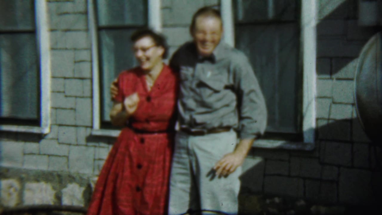 Adult woman in a red dress and adult man in gray stand smiling by windows.