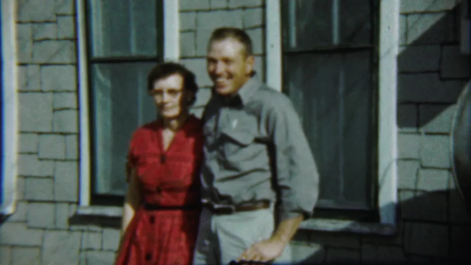 The couple remains posed outside the gray shingled house, with the woman in a red dress and the man in a gray shirt smiling toward the camera.