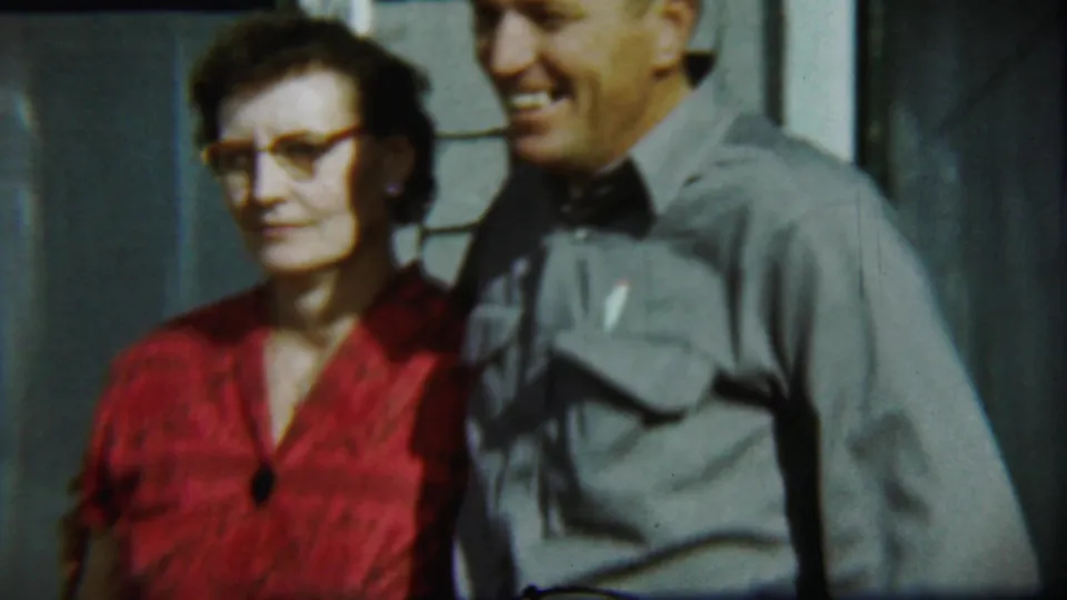 A closer 1959 home-movie view shows the woman in glasses and red dress beside the smiling man in a gray shirt against the house exterior.