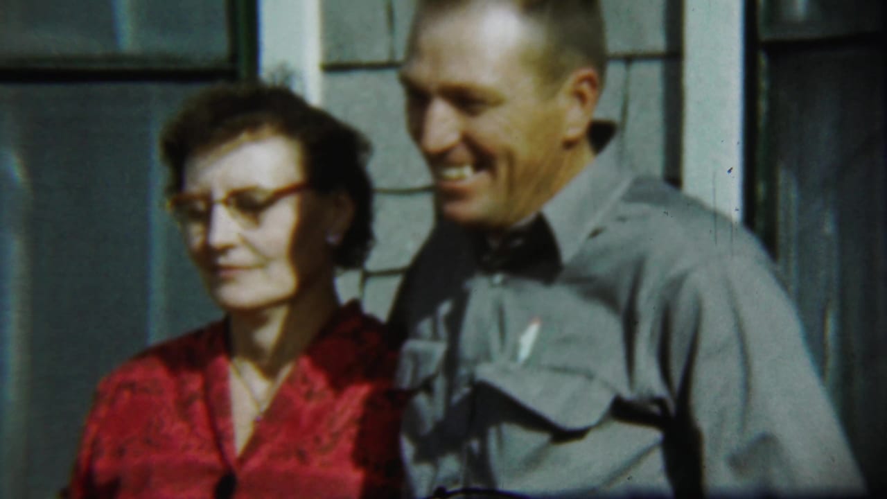 Older woman in a red dress and older man in a gray shirt stand outside a house.