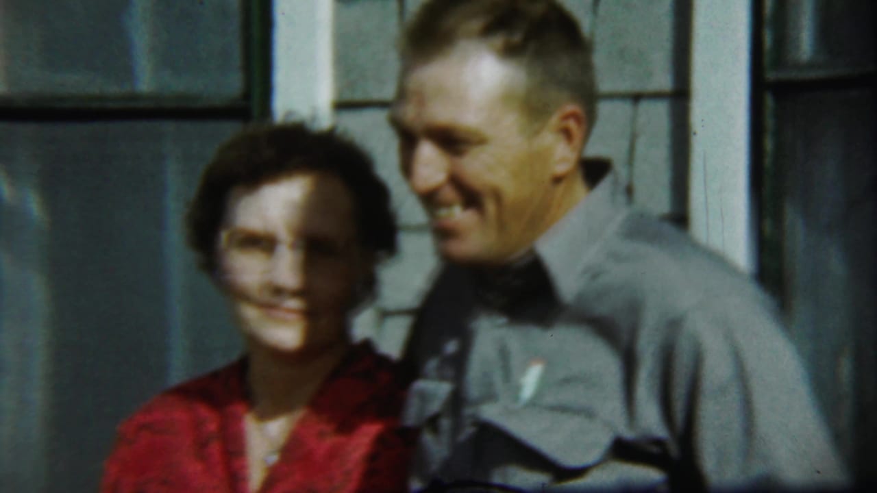 Older couple smiles beside a window on a shingled house exterior.