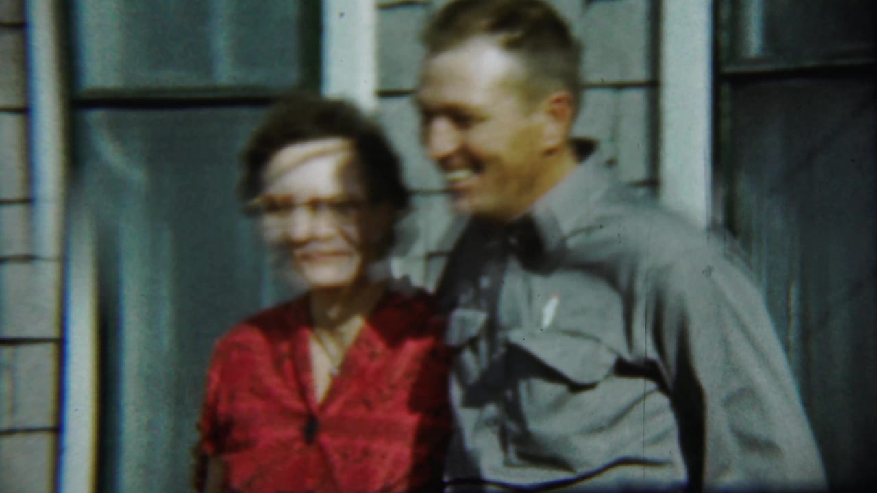 Woman in a red dress and man in a gray shirt standing outside a shingled house.