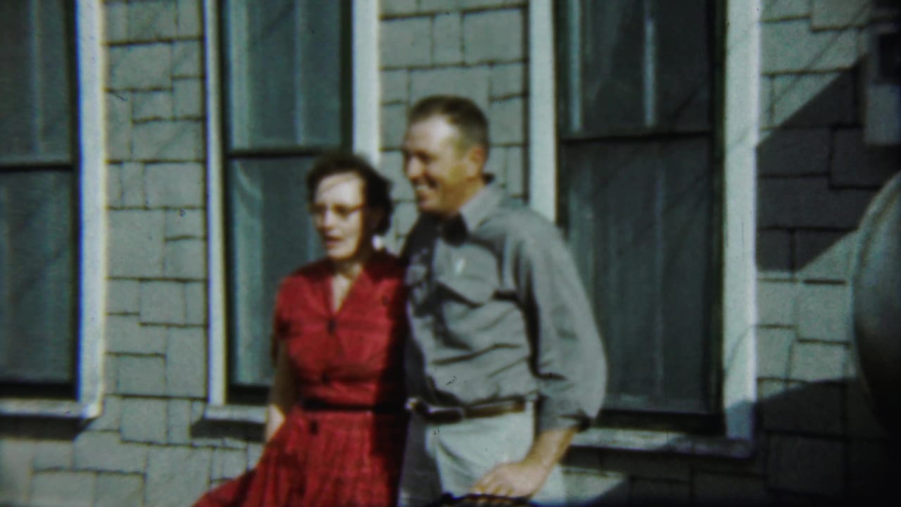 Woman in red and man in gray standing farther back beside tall house windows.