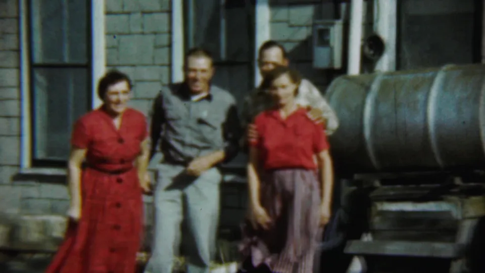 A small group gathers outside the Chadron residence, with adults in red clothing, a plaid shirt, and a large cylindrical tank nearby.