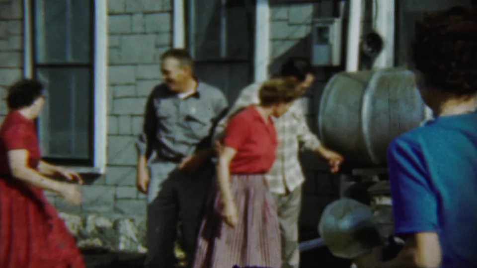 Adults interact casually in the yard beside the shingled Chadron house, with one person seen from behind at the frame edge.