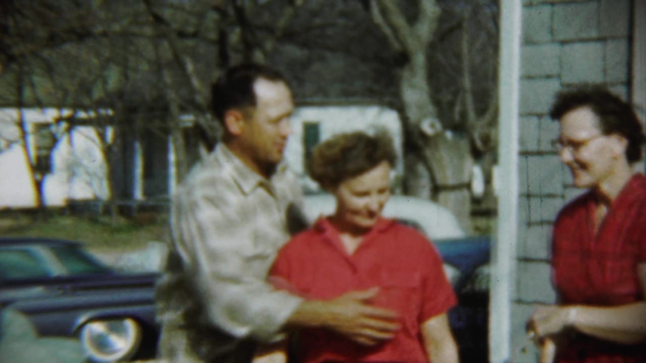 Adults standing outside a shingled house while a man embraces a woman in red.