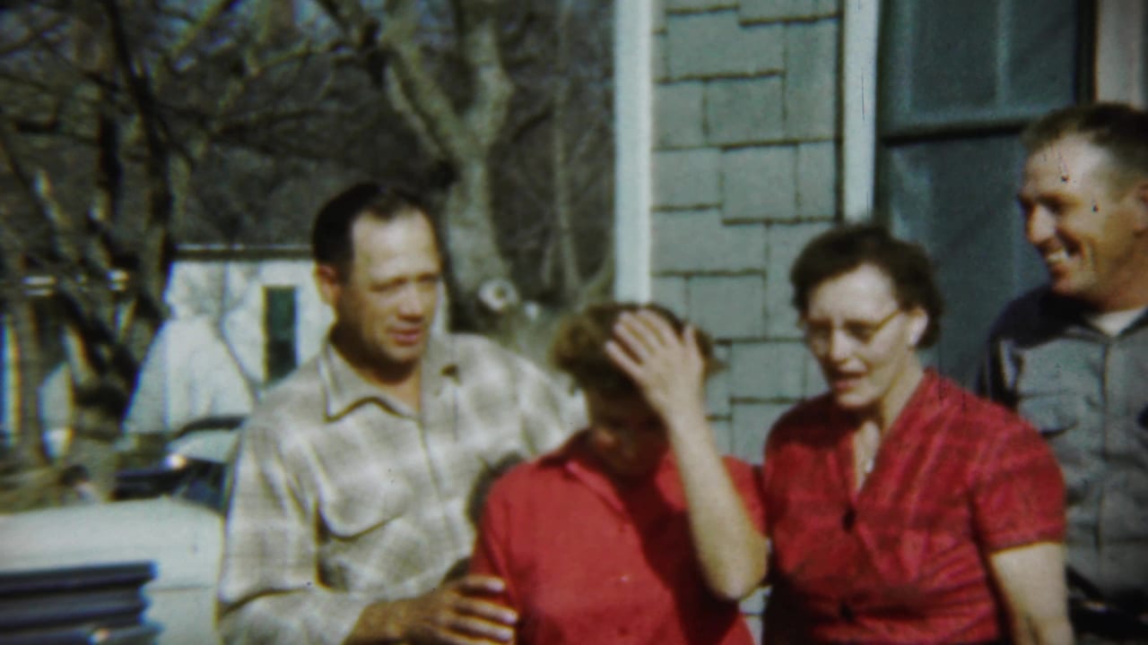 A 1959 Chadron family group stands near a house as one woman in red covers her face and others smile toward the camera.