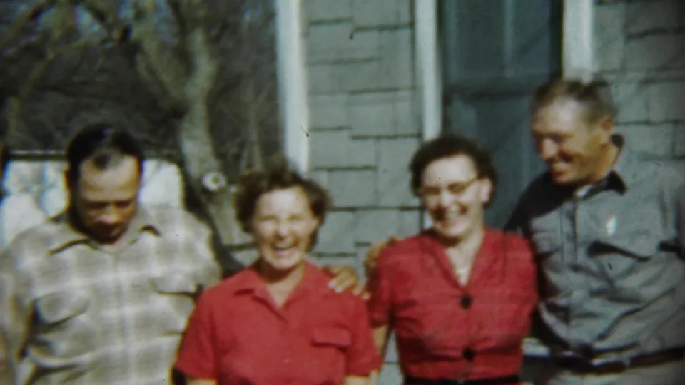 In Chadron, Nebraska in 1959, four adults laugh while posing outside a house, framed by siding, a window, and bare trees.
