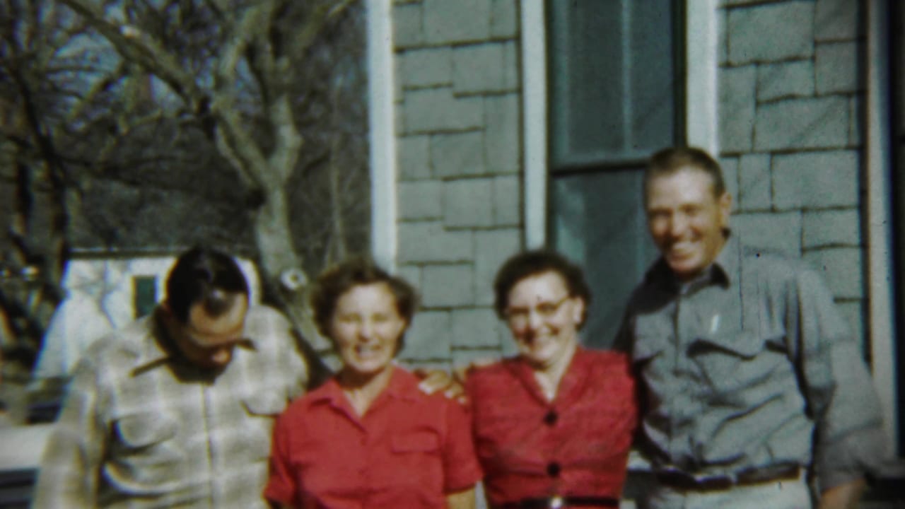 Four adults stand outside a shingled house while one man looks down and others smile.