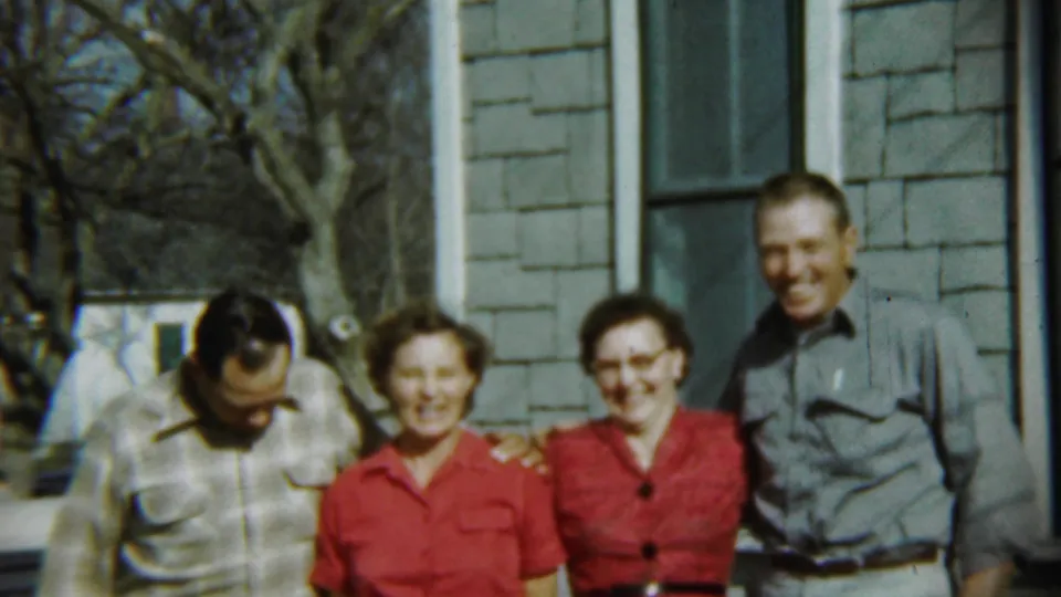 In 1959 Chadron, Nebraska, four adults pose outdoors by a shingled house, with two women in red dresses and two men in casual shirts.