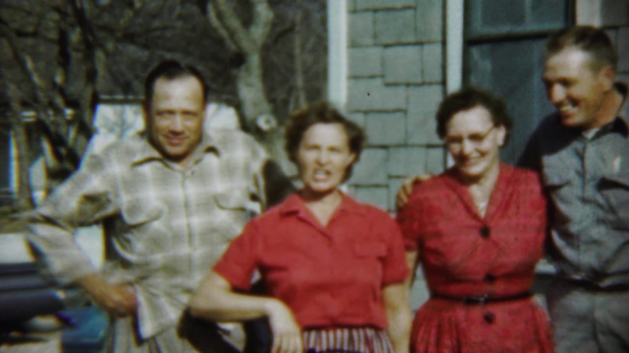 Four adults smile and laugh while standing close together outside a house.