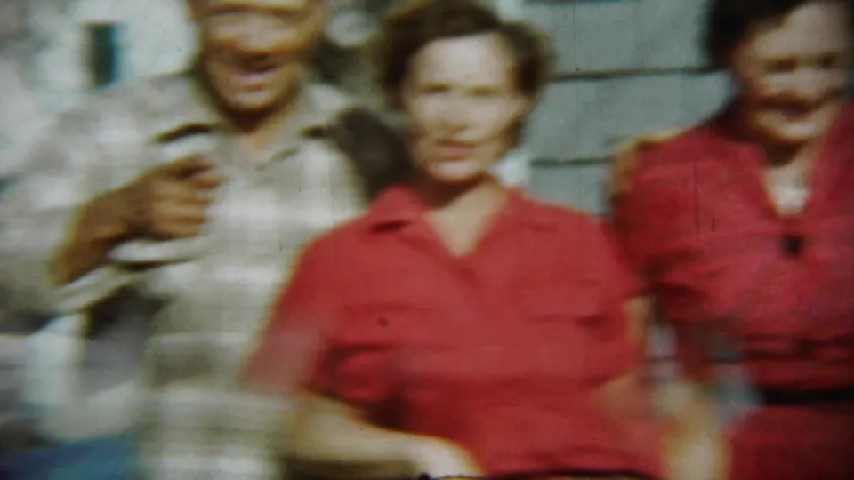 A soft-focus 8mm frame shows three adults close to the camera outside the Chadron house, with faces and red clothing visible.