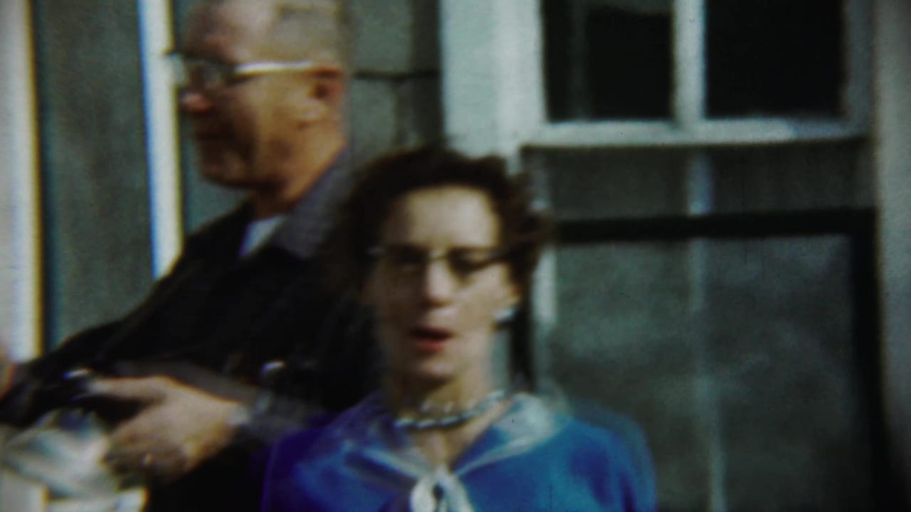 Man and woman standing on a porch, with the woman in glasses and a blue dress.