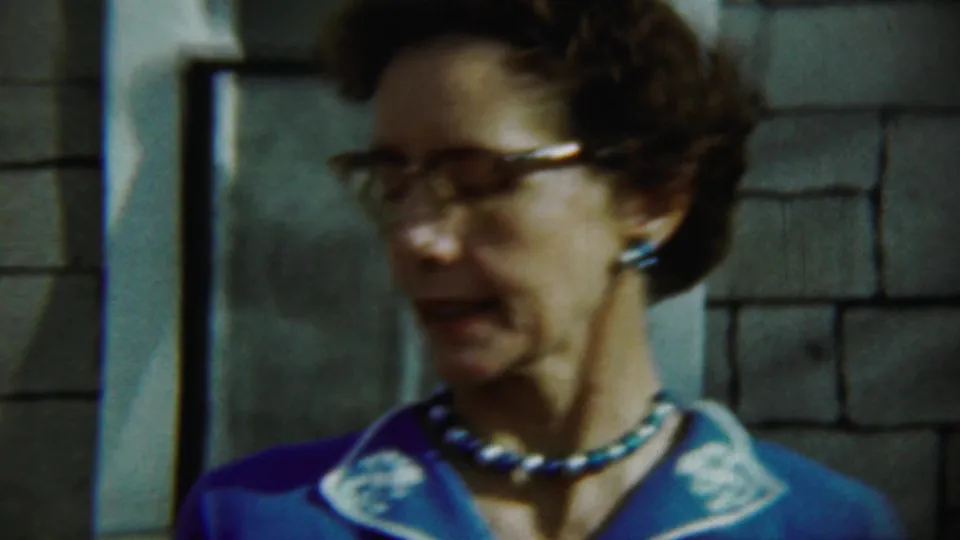 Close home-movie frame of the woman in glasses wearing a blue dress with white trim and a beaded necklace.
