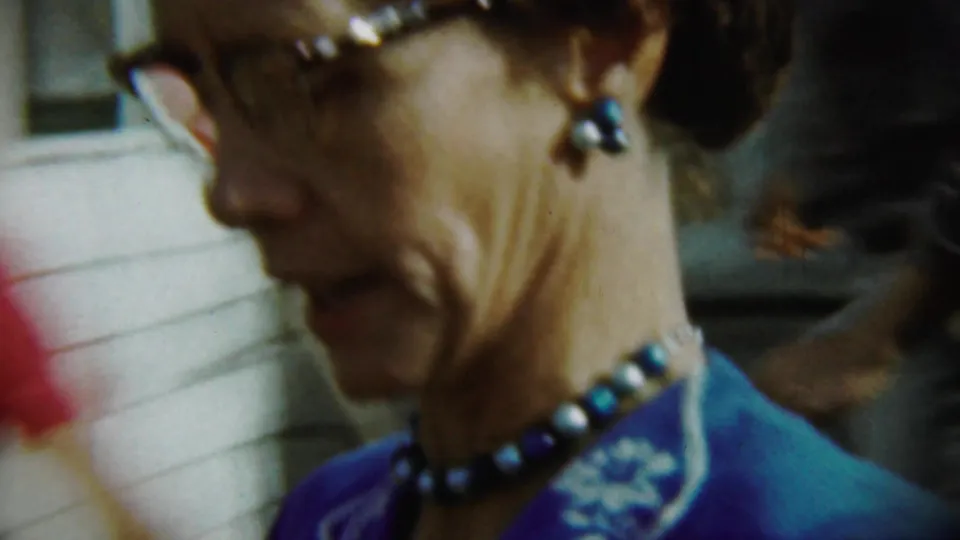 Side close-up of the woman in glasses, blue dress, earrings, and beaded necklace in the 1959 Chadron home movie.