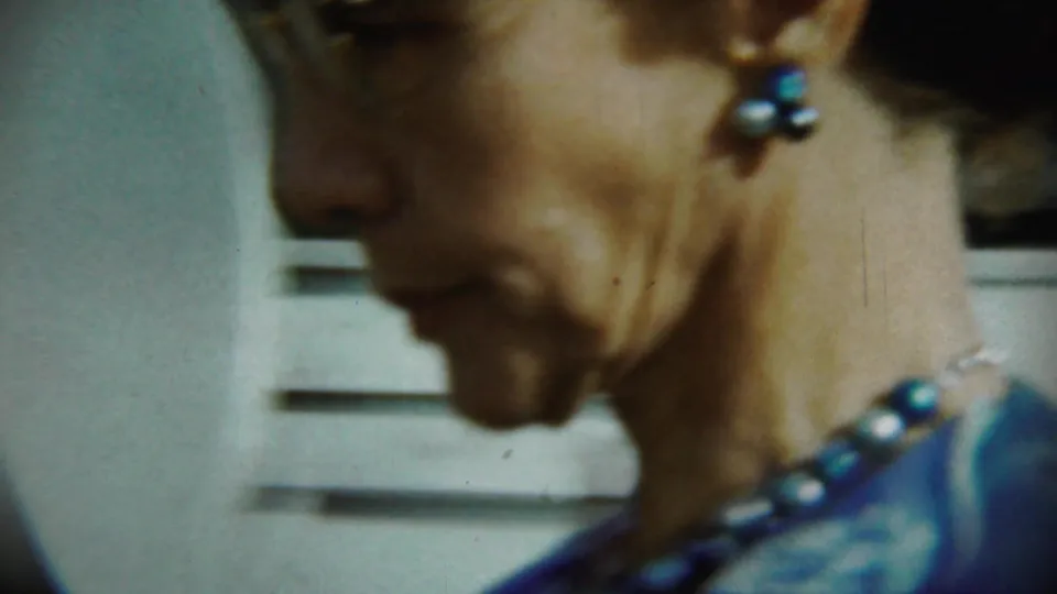 Close 8mm frame of a young woman in profile, wearing blue earrings and necklace, with a house exterior blurred behind her.