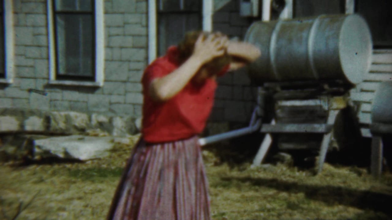 Young woman in a red shirt and striped skirt covers her face beside a brick house and metal tank.