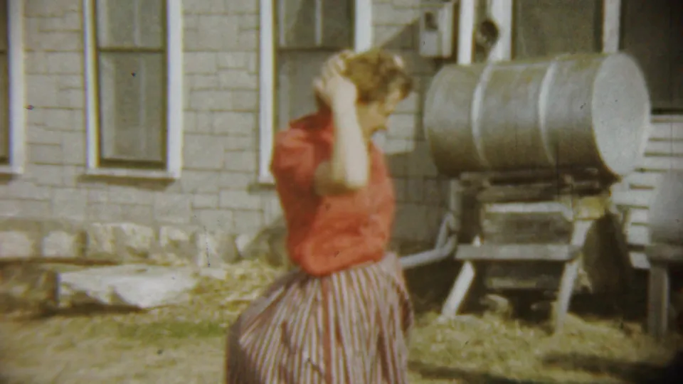 The woman adjusts her hair in a Chadron, Nebraska backyard, with a brick wall, windows, and metal yard tank behind her.