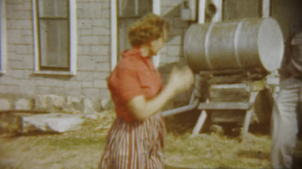 The woman stands in profile in the yard near the metal tank, while another adult figure is partly visible at the frame edge.