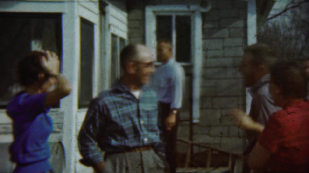 Adults gathered outside a house, with a man in a blue plaid shirt and a woman adjusting her hair.