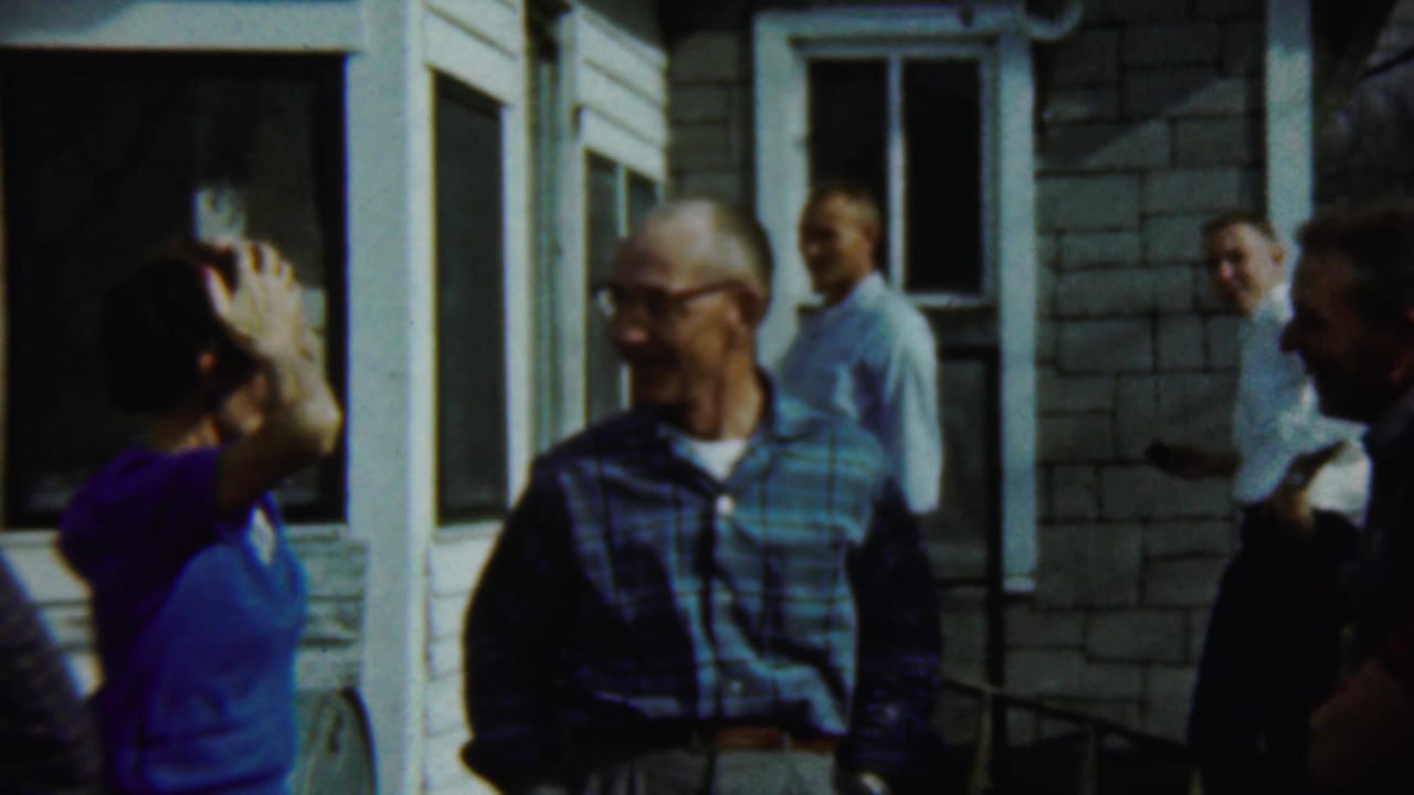 A man in a blue plaid shirt smiles while other adults stand near the porch and doorway.