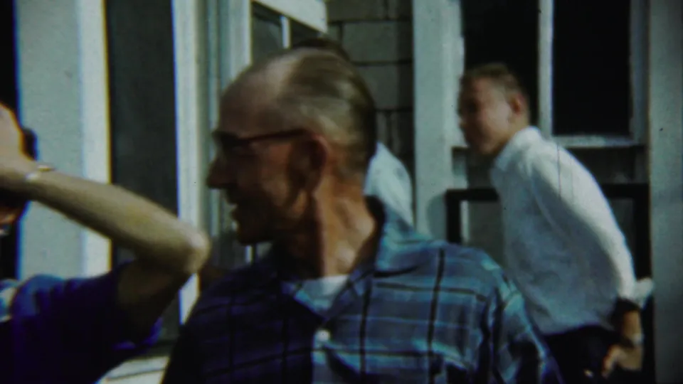 Close 1959 Chadron home-movie view of adults on a porch, with the man in glasses turned in profile beside white trim.