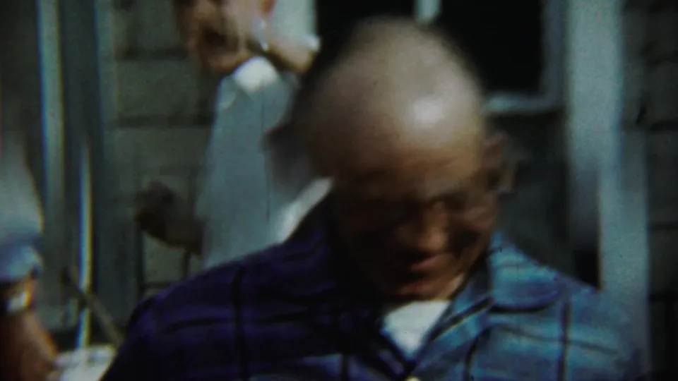 Blurred 1959 Chadron frame of a man in glasses moving close to the handheld camera beside the porch.
